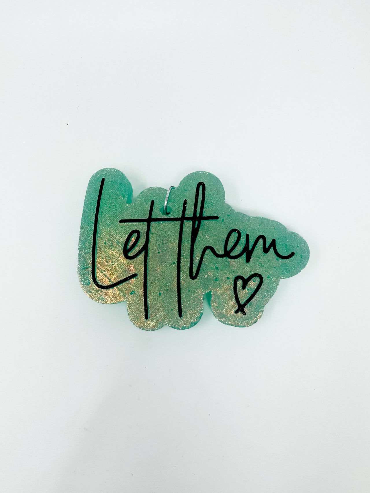 A handmade car air freshener, or freshie, featuring a shimmering sage green background with a golden glitter undertone, shown against a solid white background. The script text "Let them" is printed across the front in black cursive alongside a small black hand-drawn heart.