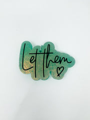 A handmade car air freshener, or freshie, featuring a shimmering sage green background with a golden glitter undertone, shown against a solid white background. The script text "Let them" is printed across the front in black cursive alongside a small black hand-drawn heart.