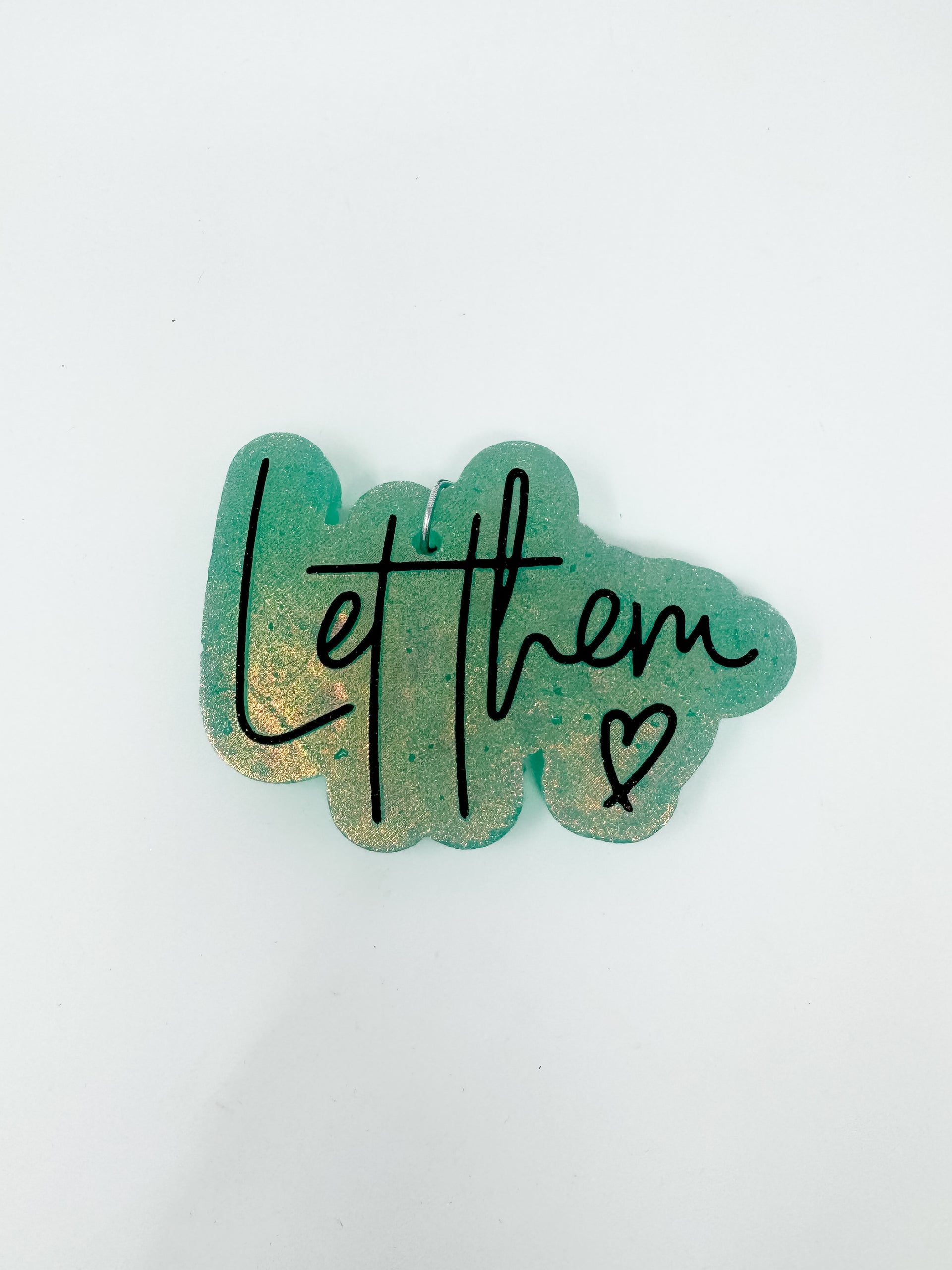 A handmade car air freshener, or freshie, featuring a shimmering sage green background with a golden glitter undertone, shown against a solid white background. The script text "Let them" is printed across the front in black cursive alongside a small black hand-drawn heart.