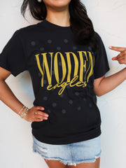 A close-up shot of a person wearing a black crewneck t-shirt with a subtle, all-over dark gray polka dot pattern. The shirt features the word "WODEN" printed in tall gold block letters with "eagles" written underneath in a matching gold cursive script font. The person is pointing to the graphic on the shirt and accessorized with layered bracelets, pairing the shirt with light-wash denim shorts.
