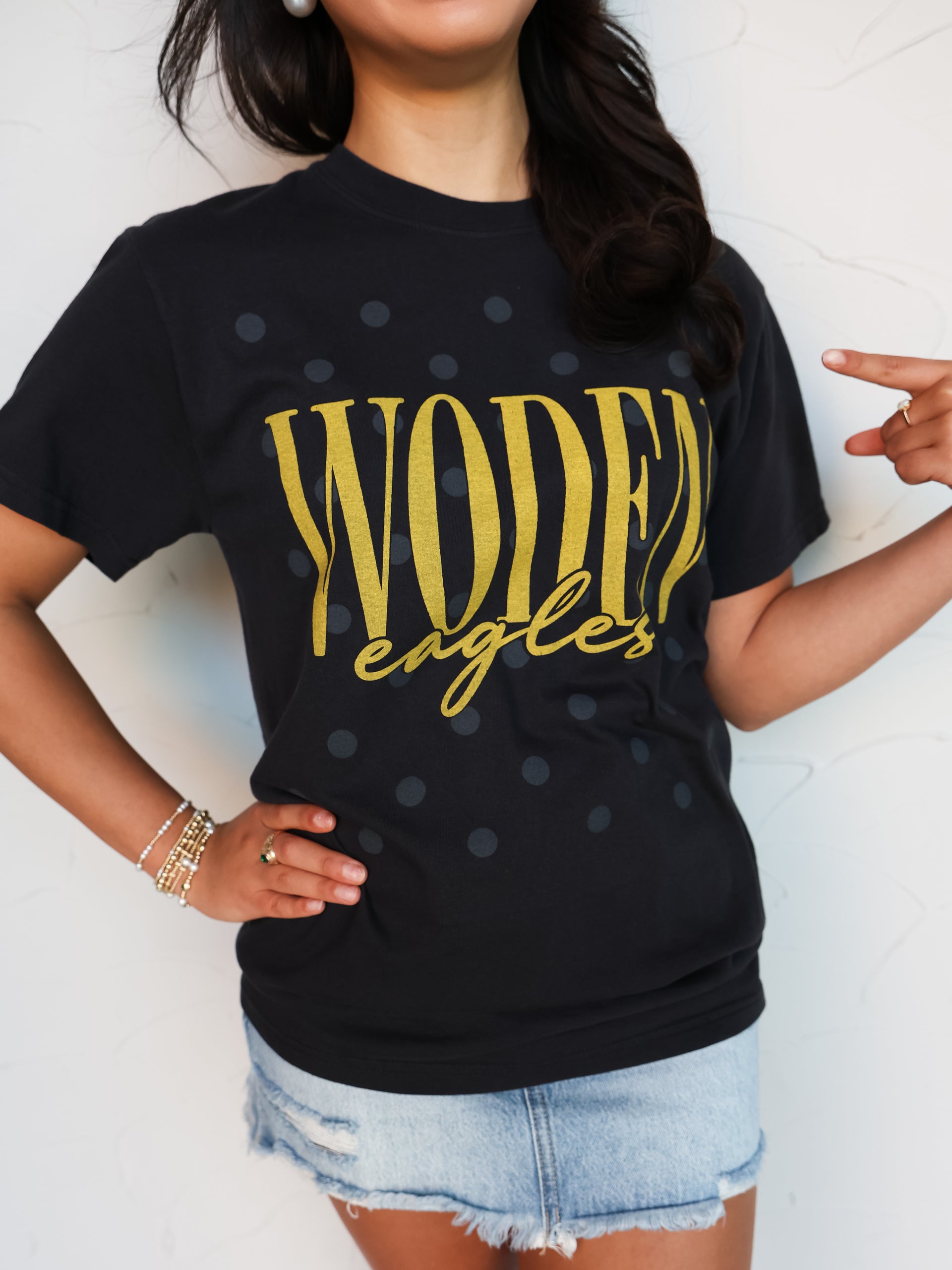 A close-up shot of a person wearing a black crewneck t-shirt with a subtle, all-over dark gray polka dot pattern. The shirt features the word "WODEN" printed in tall gold block letters with "eagles" written underneath in a matching gold cursive script font. The person is pointing to the graphic on the shirt and accessorized with layered bracelets, pairing the shirt with light-wash denim shorts.