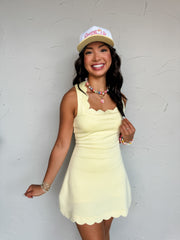 A sleeveless, light yellow active dress made from a blend of 49% polyester, 45% rayon, and 6% spandex. The dress features a distinctive scalloped square neckline, wide tank-style shoulder straps, and a fitted bodice that shapes into a slight A-line mini skirt. The bottom hem of the skirt is finished with matching scalloped trim detailing, and the design is fully lined with hidden, built-in shorts underneath.