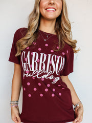 A close-up shot of a smiling person wearing a maroon crewneck t-shirt with an all-over pink polka dot pattern. The shirt features the word "GARRISON" printed in tall white block letters with "bulldogs" written underneath in a white cursive script font. The person is accessorized with silver layered necklaces and beaded bracelets.