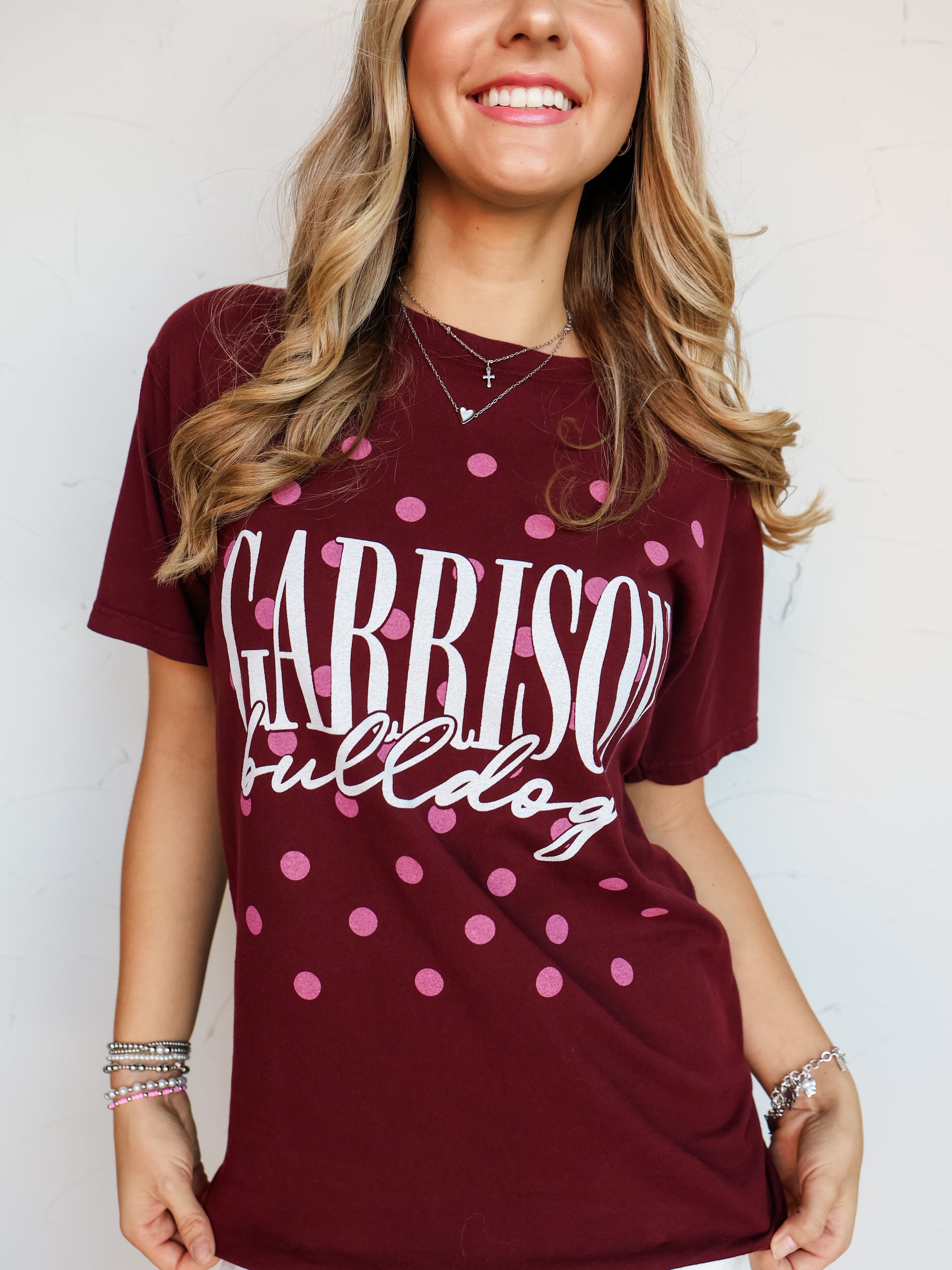 A close-up shot of a smiling person wearing a maroon crewneck t-shirt with an all-over pink polka dot pattern. The shirt features the word "GARRISON" printed in tall white block letters with "bulldogs" written underneath in a white cursive script font. The person is accessorized with silver layered necklaces and beaded bracelets.