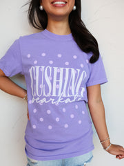 A close-up shot of a smiling person wearing a purple crewneck t-shirt with an all-over lavender polka dot pattern. The shirt features the word "CUSHING" printed in tall white block letters with "bearcats" written underneath in a white cursive script font. The person is accessorized with large white pearl earrings and beaded bracelets, pairing the shirt with light-wash denim shorts.
