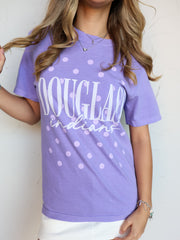 A close-up shot of a person wearing a purple crewneck t-shirt with an all-over lavender polka dot pattern. The shirt features the word "DOUGLASS" printed in tall white block letters with "indians" written underneath in a white cursive script font. The person is accessorized with silver layered necklaces and bracelets, pairing the shirt with white bottoms.