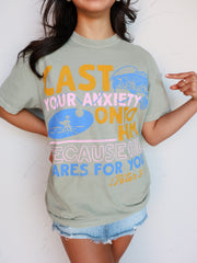 A close-up shot of a smiling person wearing a sage green graphic tee with the scripture "Cast your anxiety onto Him because He cares for you 1 Peter 5:7." The text is printed in a colorful, slightly distressed font with mustard yellow, pink, and blue colors, accompanied by illustrations of a fisherman and a fish. The person is pointing at the design and wearing light wash distressed denim shorts.