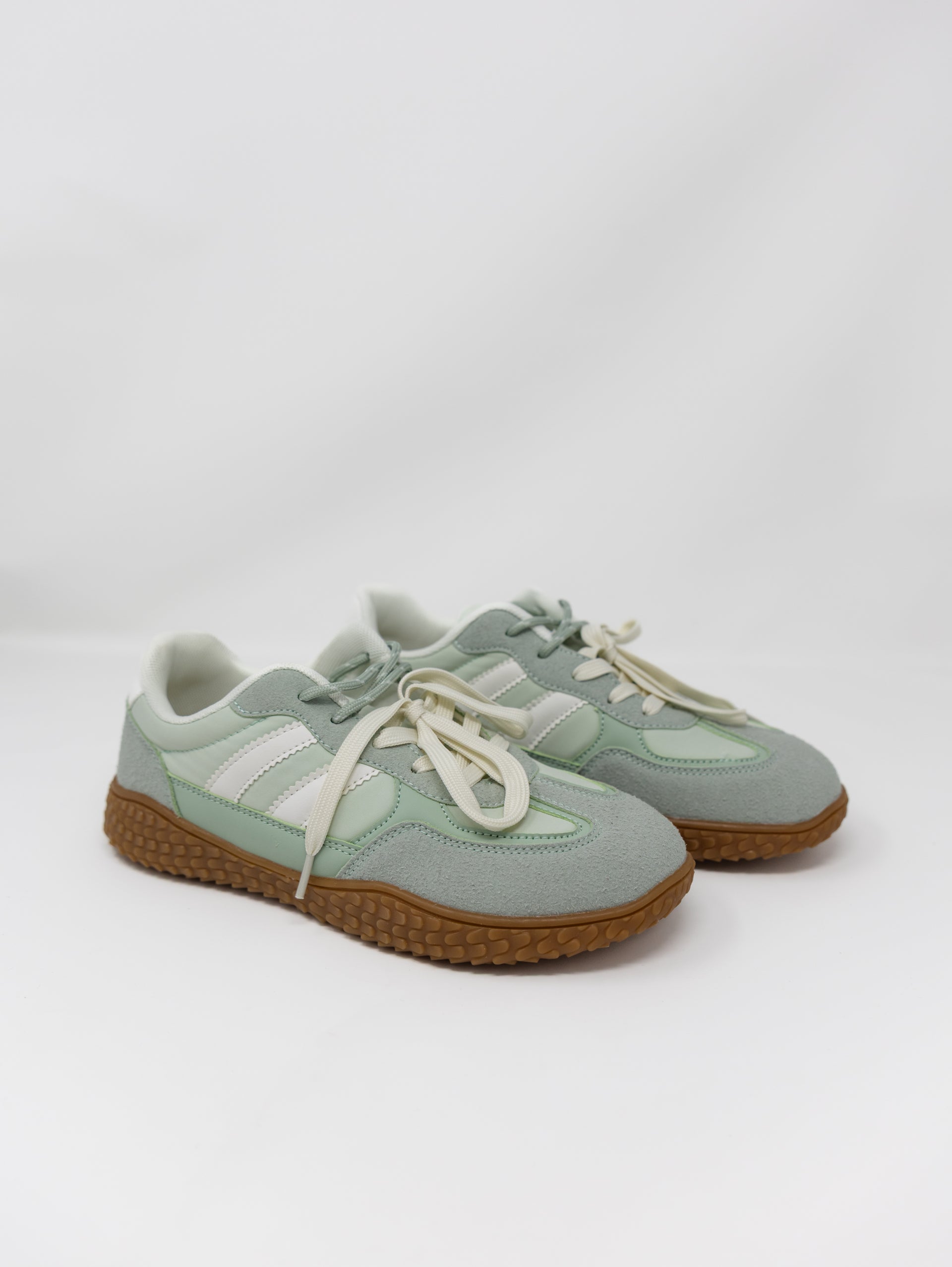 A pair of low-top sneakers in a muted sage green colorway, featuring cream-colored laces, white side stripes, and brown textured rubber soles, photographed on a light gray studio background.