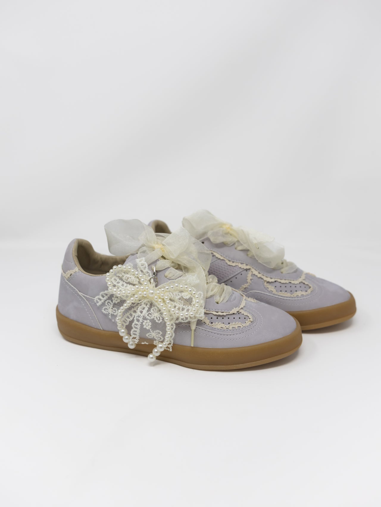 A pair of light lavender suede sneakers decorated with cream lace bows and ruffles, featuring tan rubber soles, photographed on a seamless white studio background.