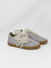 A pair of light lavender suede sneakers decorated with cream lace bows and ruffles, featuring tan rubber soles, photographed on a seamless white studio background.