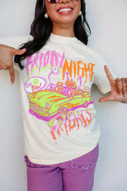 A woman wearing a cream-colored graphic t-shirt with a colorful 'Friday Night' Halloween design, paired with purple pants, posing against a white wall.