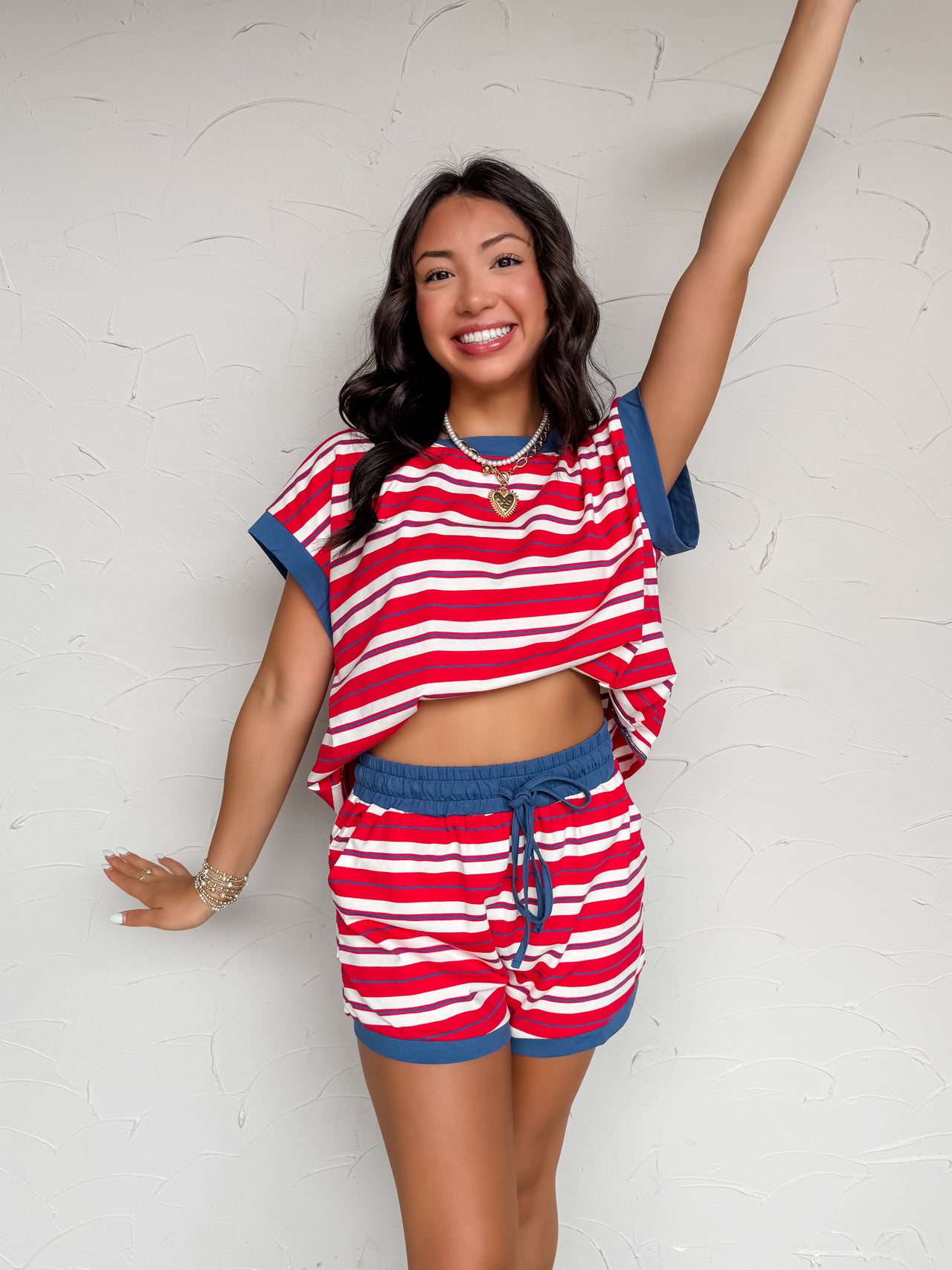 A two-piece casual lounge set featuring a vibrant red, white, and thin blue horizontal stripe pattern, crafted from a soft knit blend of 95% cotton and 5% spandex. The set includes an oversized, relaxed-fit short-sleeve top with a solid blue crew neckline and matching blue folded sleeve cuffs. The coordinating shorts have an elastic waistband with a blue drawstring and matching solid blue fabric trim along the bottom hems.