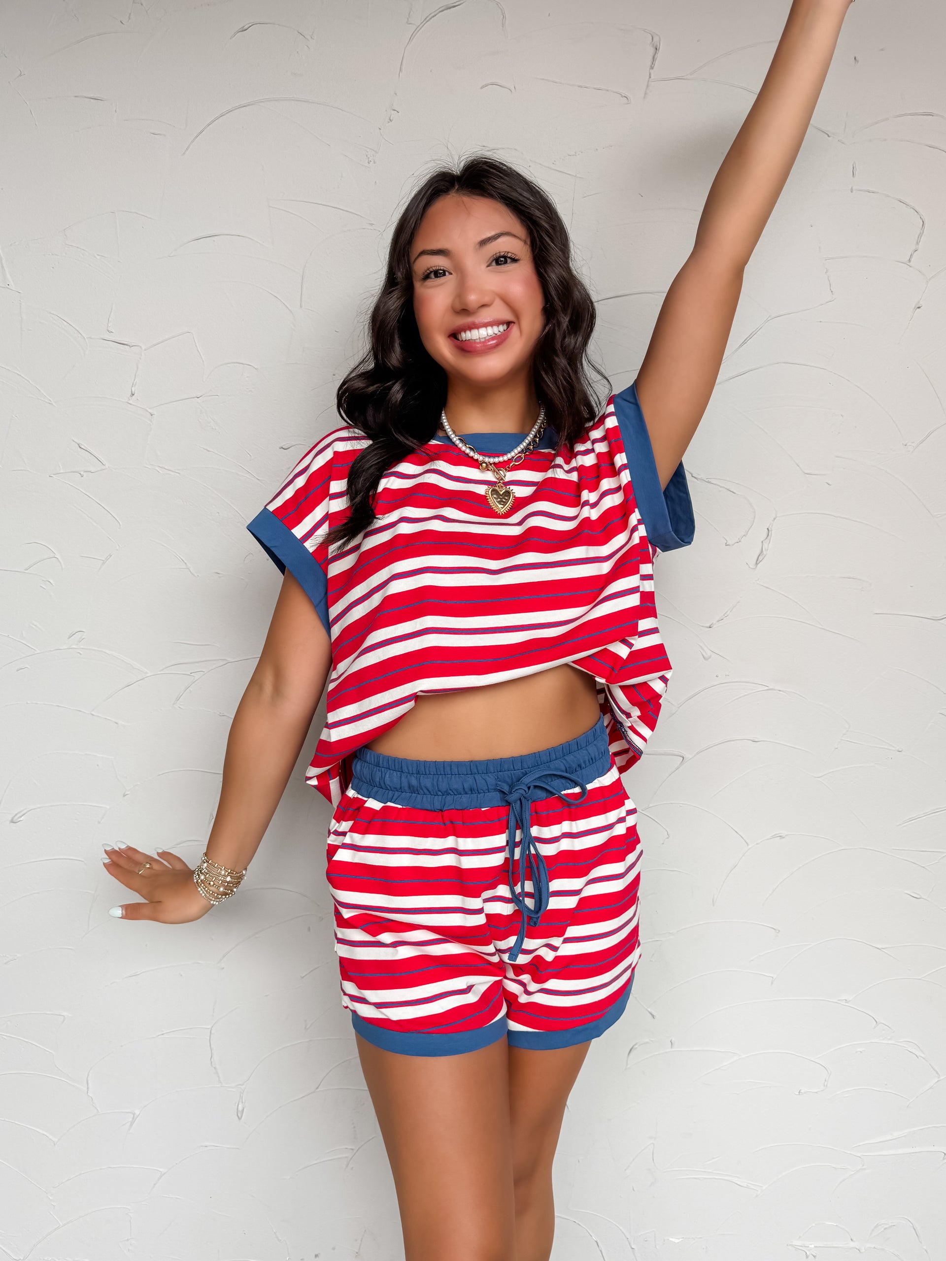 A two-piece casual lounge set featuring a vibrant red, white, and thin blue horizontal stripe pattern, crafted from a soft knit blend of 95% cotton and 5% spandex. The set includes an oversized, relaxed-fit short-sleeve top with a solid blue crew neckline and matching blue folded sleeve cuffs. The coordinating shorts have an elastic waistband with a blue drawstring and matching solid blue fabric trim along the bottom hems.