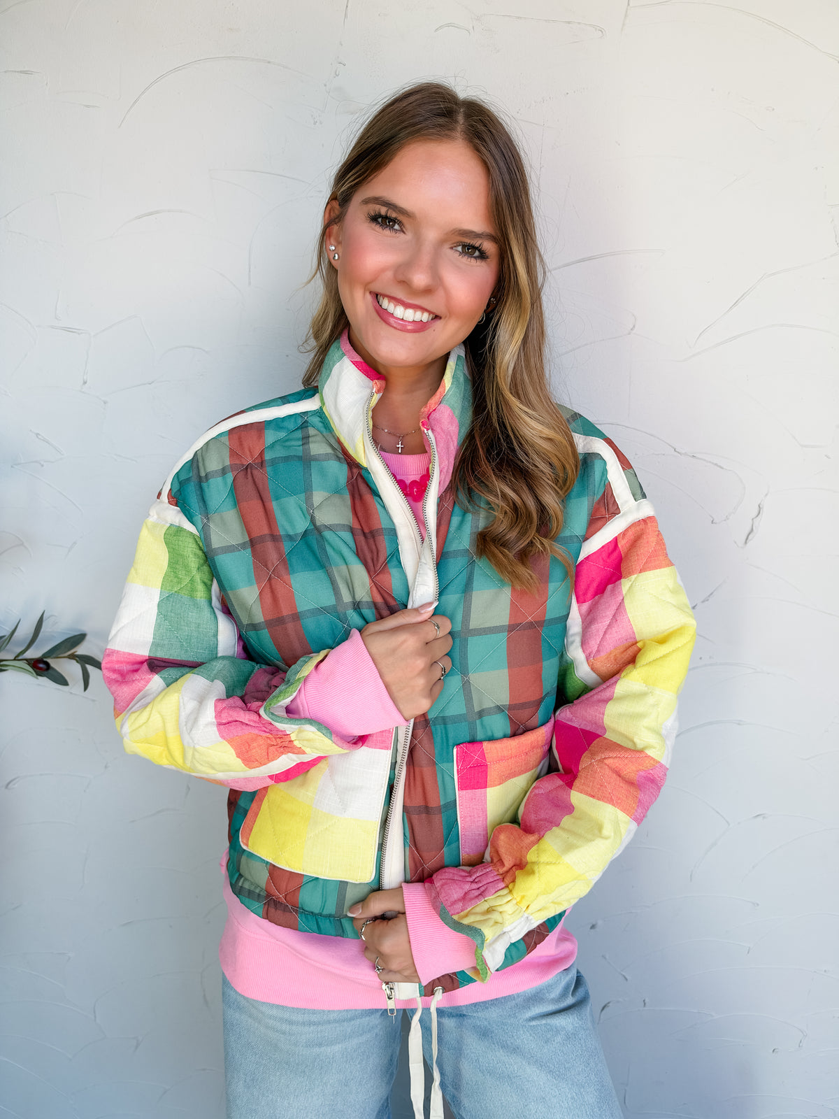 Catie modeling the Something Cute Quilted Patchwork Puffer Jacket by J. NNA, made of 100% polyester, with diamond quilting, a teal and rust plaid back, contrasting pink, yellow, and cream plaid sleeves, and a stand collar, styled with light-wash jeans.