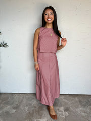 A full-length photograph of a smiling brunette woman with long straight dark hair holding a strand of her hair while looking to the side against a textured off-white wall next to a potted olive plant. She is wearing a pink and brown striped two-piece set made from an 80% polyester and 20% cotton blend, featuring a sleeveless high-neck cropped top with vertical stripes and a matching high-waisted ankle-length maxi skirt with diagonal chevron stripes, styled with cognac brown leather ballet flats, a gold circ