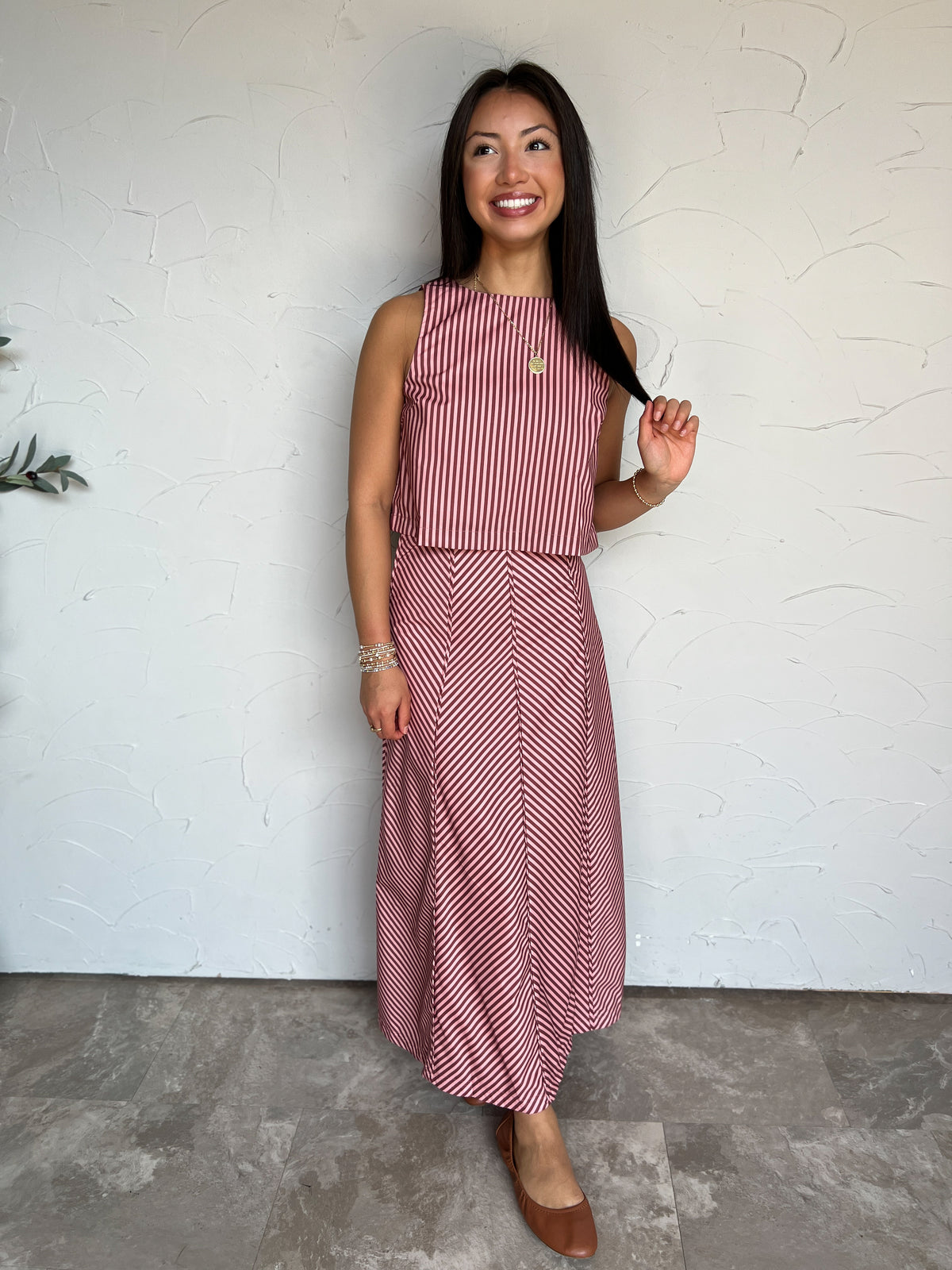 A full-length photograph of a smiling brunette woman with long straight dark hair holding a strand of her hair while looking to the side against a textured off-white wall next to a potted olive plant. She is wearing a pink and brown striped two-piece set made from an 80% polyester and 20% cotton blend, featuring a sleeveless high-neck cropped top with vertical stripes and a matching high-waisted ankle-length maxi skirt with diagonal chevron stripes, styled with cognac brown leather ballet flats, a gold circ