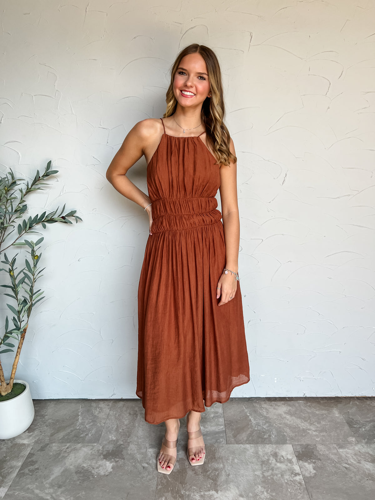 A full-length photograph of a smiling young woman with long wavy blonde hair posing with one hand on her hip against a textured off-white wall next to a potted olive plant. She is wearing a sleeveless rust-brown midi dress made from an 80% rayon and 20% nylon blend, featuring thin halter straps, an apron-style high neck, a wide smocked elastic waist, and a flowing pleated skirt, styled with clear double-strap slide heels, a thin silver cross necklace, a silver charm bracelet, and red nail polish.