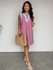 A full-length view of a woman modeling a sleeveless shift-style tunic dress made from 100% cotton. The dress features a mixed gingham check design, with a red and white gingham main body contrasted by light blue and white gingham trim along the armholes and the split V-neckline. The neckline is accented with white piped trim. She is accessorized with layered gold necklaces, bracelets, white block-heeled sandals, and is holding a woven straw tote bag with structured wooden handles.