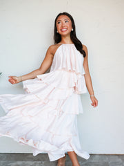 A full-length photograph of a smiling brunette woman posing against a textured white wall while holding the side of her dress. She is wearing a flowing light pink halter maxi dress made from an 80% rayon and 20% nylon blend crinkle fabric, featuring a high neckline and a tiered, ruffled skirt.