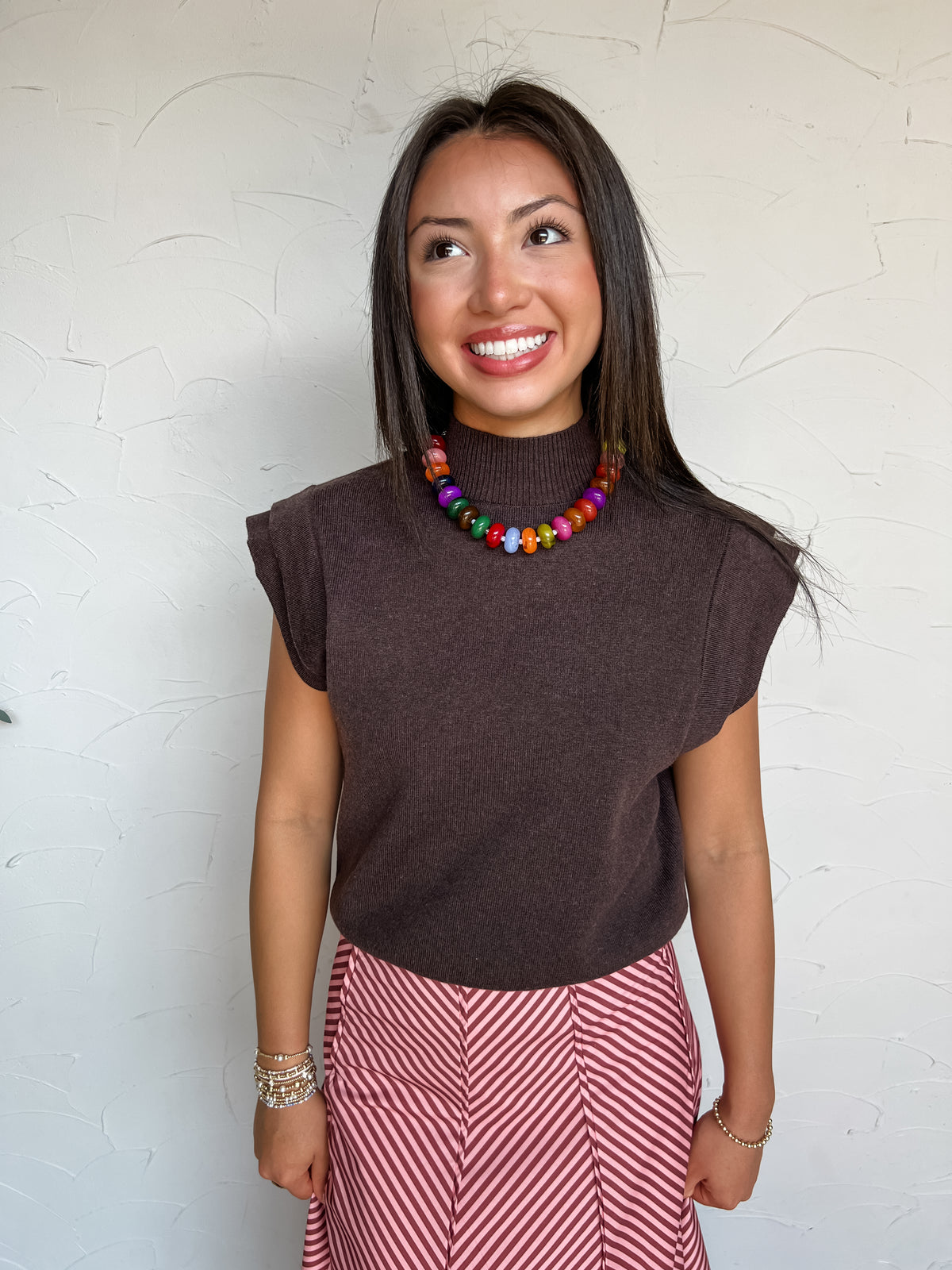 A portrait photograph of a smiling brunette woman with long straight dark hair looking upward and to the side while posing against a textured off-white wall. She is wearing a short-sleeved knit sweater top in a dark chocolate brown shade, made from a 54% polyester, 20% nylon, and 20% acrylic blend, featuring a high ribbed mock neck, cap shoulders, and a fitted waistband, paired with pink and red diagonal striped pants, styled with a colorful chunky beaded necklace and stacked gold and beaded bracelets.