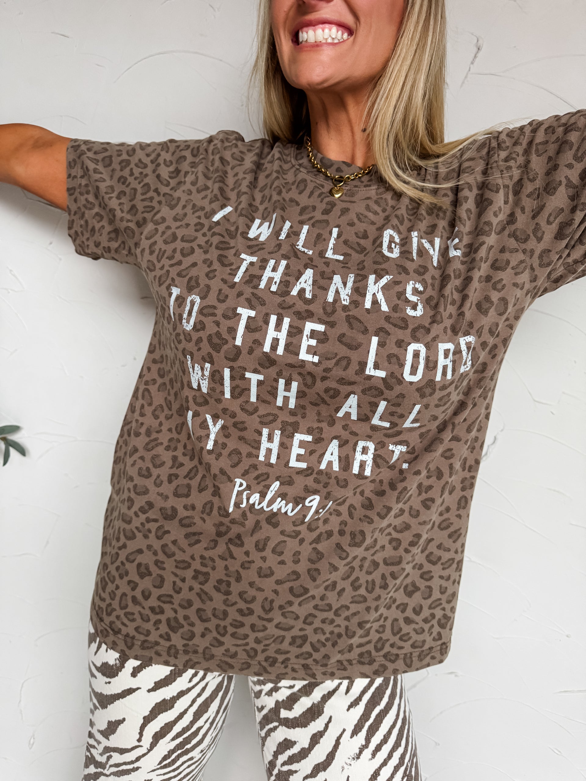 A woman wearing a brown leopard-print t-shirt with white text that reads 'I WILL GIVE THANKS TO THE LORD WITH ALL MY HEART Psalm 94' paired with zebra-print leggings, posing against a white wall.