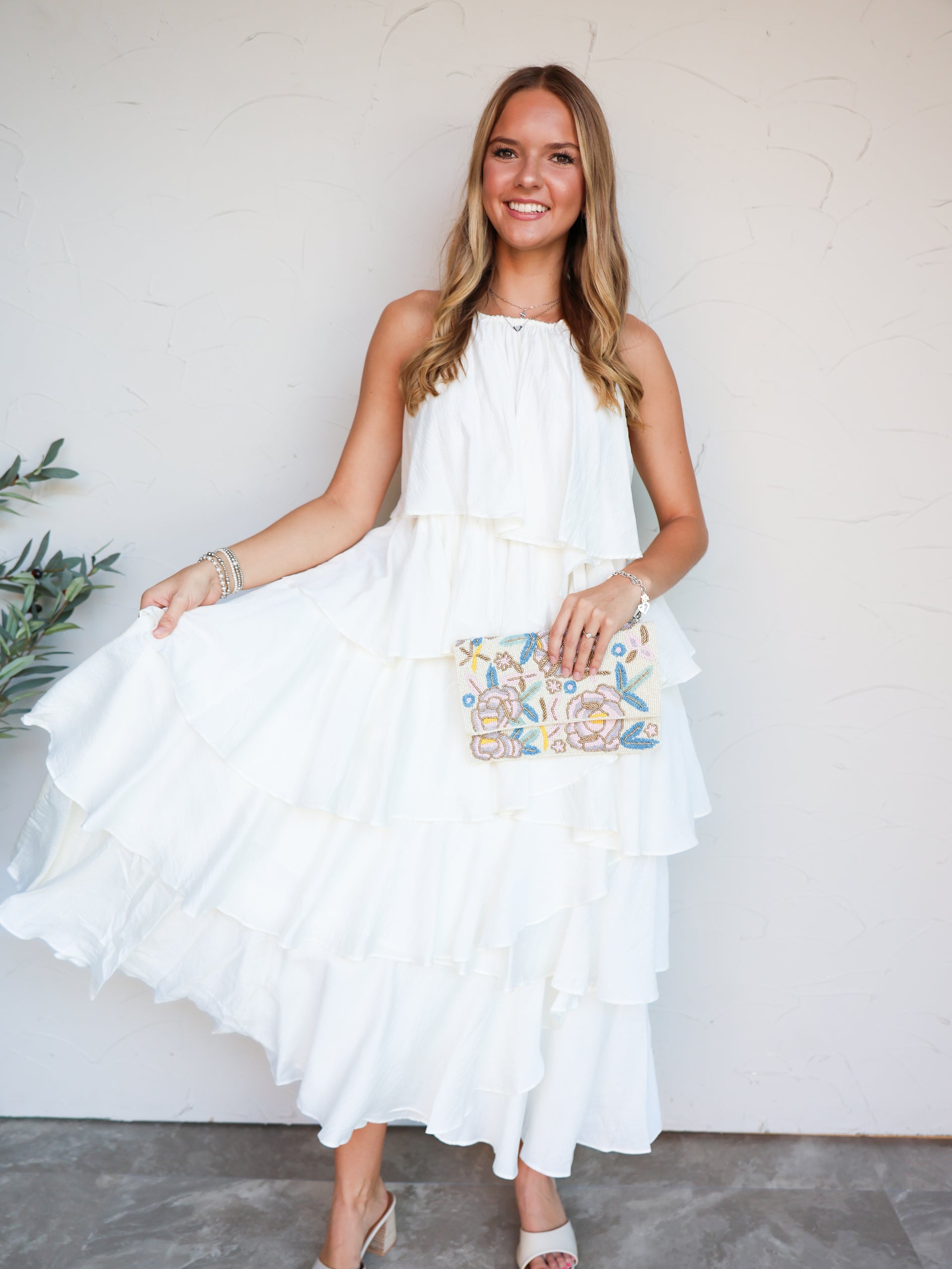A full-length photograph of a smiling blonde woman posing against a textured white wall while holding the side of her dress. She is wearing a flowing off-white halter maxi dress made from an 80% rayon and 20% nylon blend crinkle fabric, featuring a high neckline and a tiered, ruffled skirt, styled with heeled mules and a floral beaded clutch.