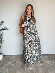A full-length photograph of a smiling young woman with long wavy blonde hair posing holding a brown shoulder bag against a textured off-white wall next to a potted olive plant. She is wearing a sleeveless floor-length maxi dress made from an 80% polyester and 20% cotton blend in a black and white zebra print, featuring a high halter neck and a flowing swing silhouette, styled with cognac brown leather boots, a brown leather shoulder bag with a chain strap, a white pearl choker necklace with a gold heart pen