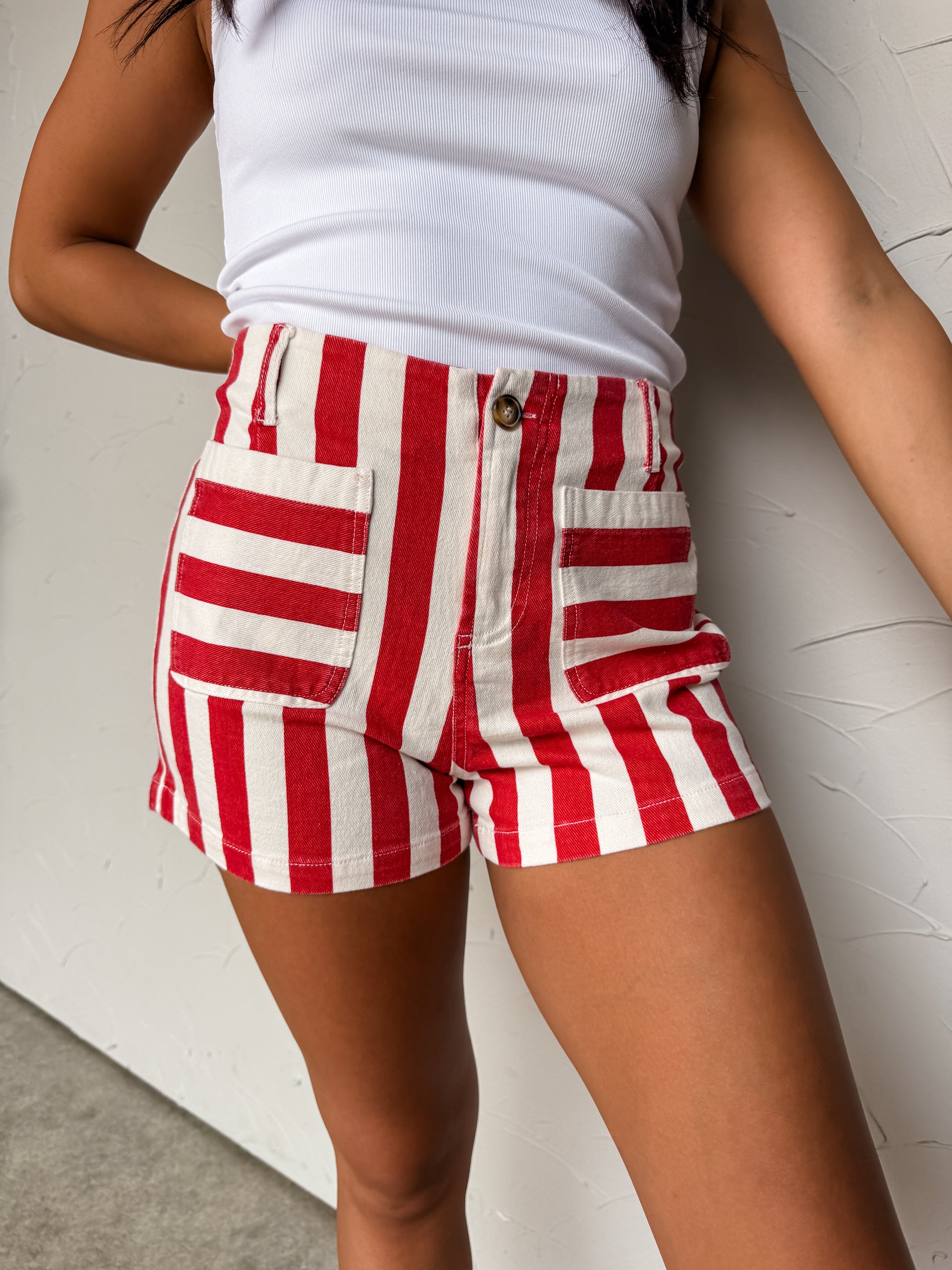 A pair of high-rise striped shorts featuring a bold red and cream-white vertical stripe pattern, made from a structured fabric blend of 97% cotton and 3% spandex. The shorts include a classic waistband with belt loops, a tortoiseshell button closure with a zip fly, and two prominent square front patch pockets designed with contrasting horizontal stripes.