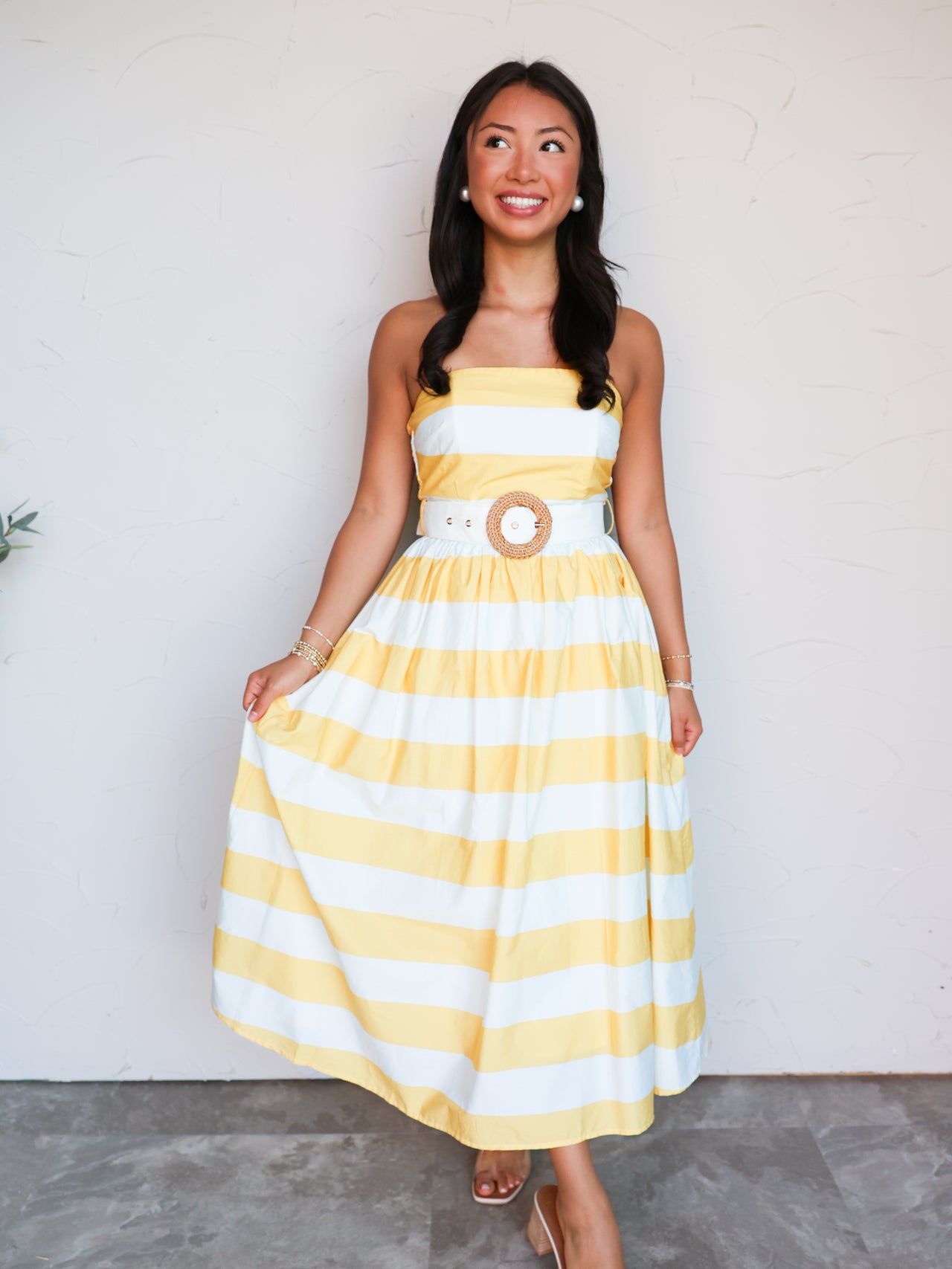 A full-length photograph of a smiling brunette woman looking off-camera and posing against a textured white wall while lightly holding her skirt. She is wearing a strapless yellow and white horizontally striped midi dress made from a 100% cotton fabric, featuring a fitted bodice, a flared pleated skirt, and a white belt with a round woven straw buckle at the waist, styled with tan heeled mules.