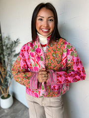 A portrait photograph of a smiling brunette woman with long straight dark hair holding the front of her jacket while posing against a textured off-white wall next to a potted olive plant. She is wearing a colorful quilted open-front jacket made with a 100% cotton shell and 100% polyester lining in a pink, red, and orange floral block-print pattern, featuring pink-and-cream striped trim along the open placket and cuffs, layered over a cream turtleneck sweater and beige pants, styled with a red beaded necklac