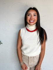 A portrait photograph of a smiling brunette woman with long straight dark hair looking upward while posing against a textured off-white wall next to a potted olive plant. She is wearing a cream off-white sleeveless knit sweater top made from a 50% acrylic, 28% polyester, and 22% nylon blend, featuring a high ribbed turtleneck collar and a straight ribbed hem, paired with light beige corduroy pants, styled with a vibrant red chunky glass bead necklace, stacked gold and pearl beaded bracelets, and a gold bead