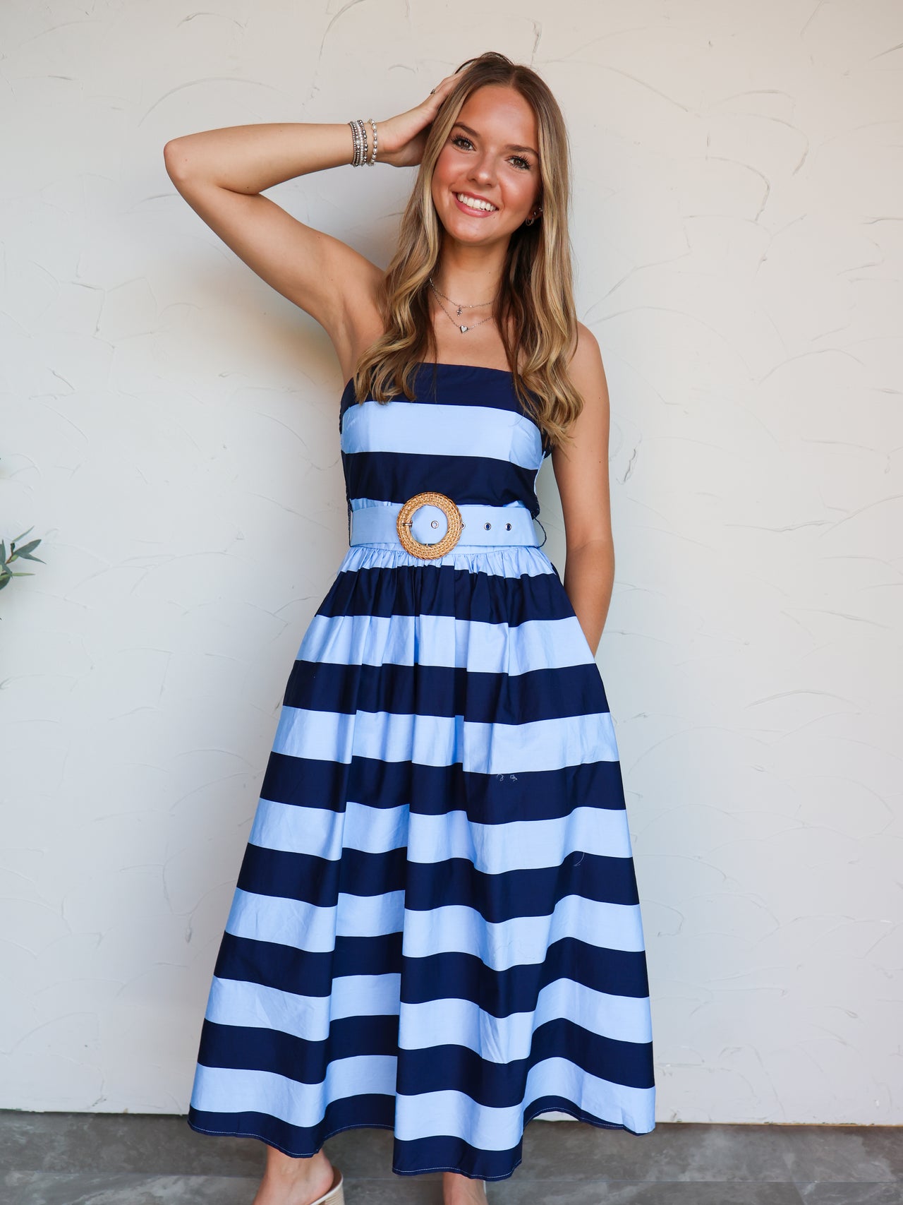 A woman modeling a strapless midi dress with horizontal navy blue and light blue stripes, featuring a matching fabric belt with a round wooden medallion, standing against a textured off-white wall.