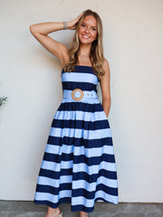 A woman modeling a strapless midi dress with horizontal navy blue and light blue stripes, featuring a matching fabric belt with a round wooden medallion, standing against a textured off-white wall.