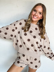 A woman wearing a beige and brown polka dot lounge set, featuring a cropped sweatshirt and matching shorts, posing against a white textured wall. 60% Cotton 40% Polyester 