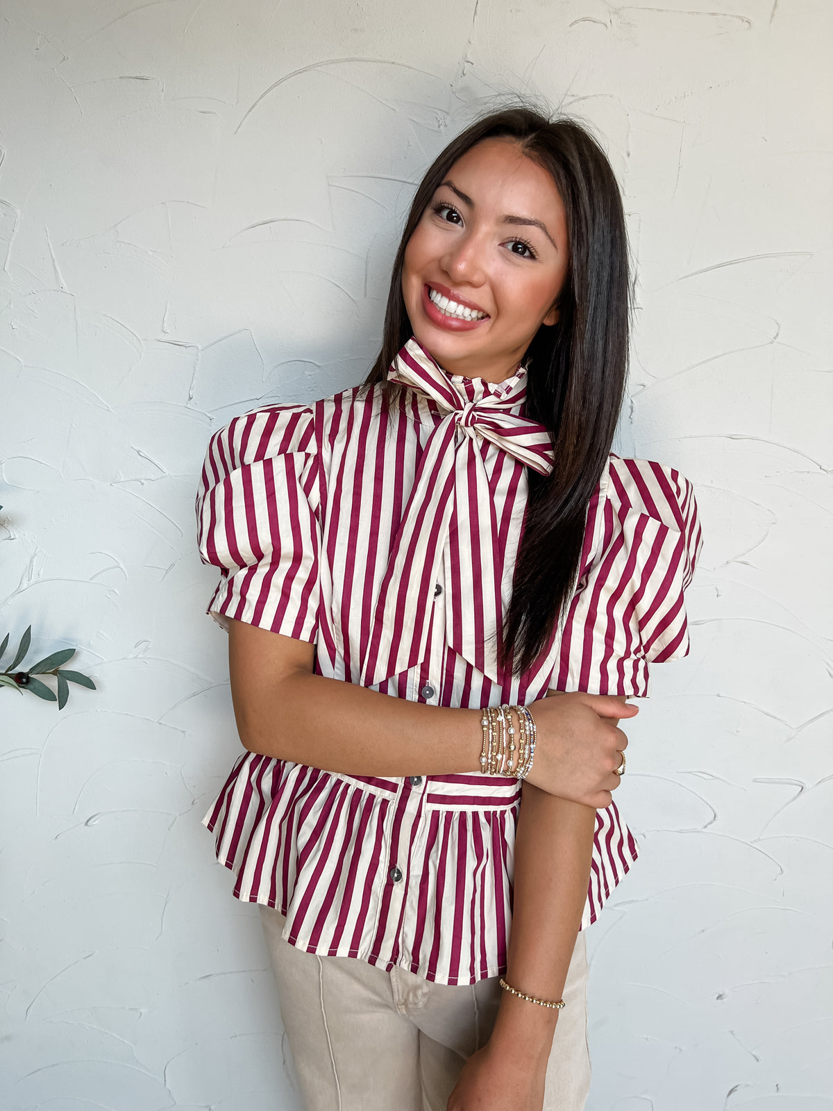 A portrait photograph of a smiling brunette woman with long straight dark hair posing with her arms crossed against a textured off-white wall next to a potted olive plant. She is wearing a short-sleeved 100% cotton blouse with vertical cream and burgundy red stripes, featuring a high ruffled collar with a statement neck bow tie, puffed short sleeves, grey button-down closures, and a fitted waist with a peplum hem, paired with light beige jeans, styled with stacked gold, pearl, and beaded bracelets.