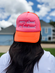 Person wearing a pink and orange cap with 'sorry about my husband' text, outdoors with houses in the background.