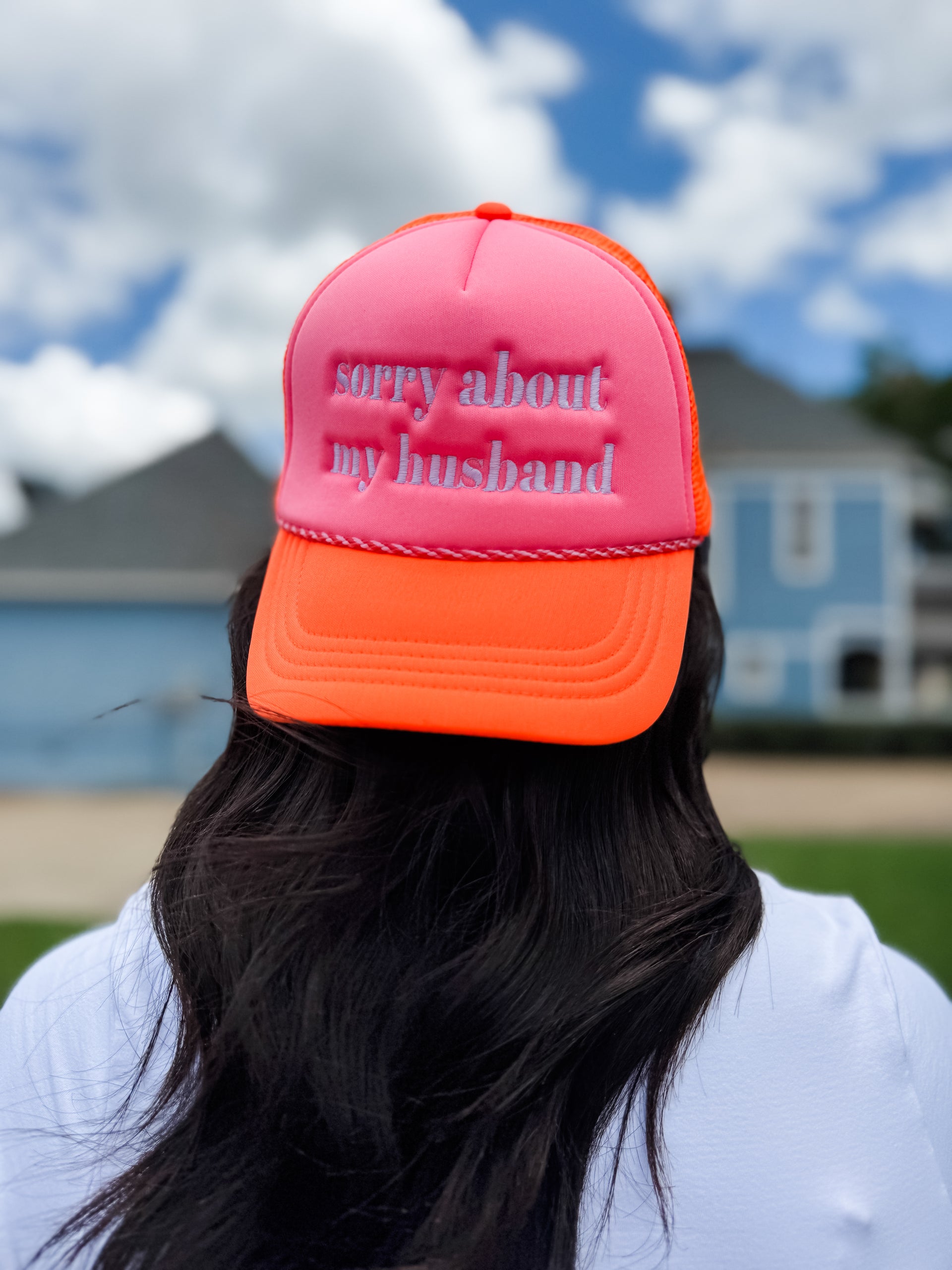 Person wearing a pink and orange cap with 'sorry about my husband' text, outdoors with houses in the background.