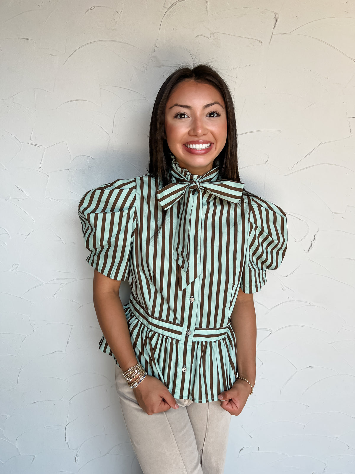 A portrait photograph of a smiling brunette woman with long straight dark hair posing with her hands in her pockets against a textured off-white wall. She is wearing a short-sleeved 100% cotton blouse with vertical mint green and dark brown stripes, featuring a high ruffled collar tied with a prominent bow, dramatic puff sleeves, a button-down front, and a seam-accented peplum hem, paired with beige pants, styled with stacked gold and pearl beaded bracelets.