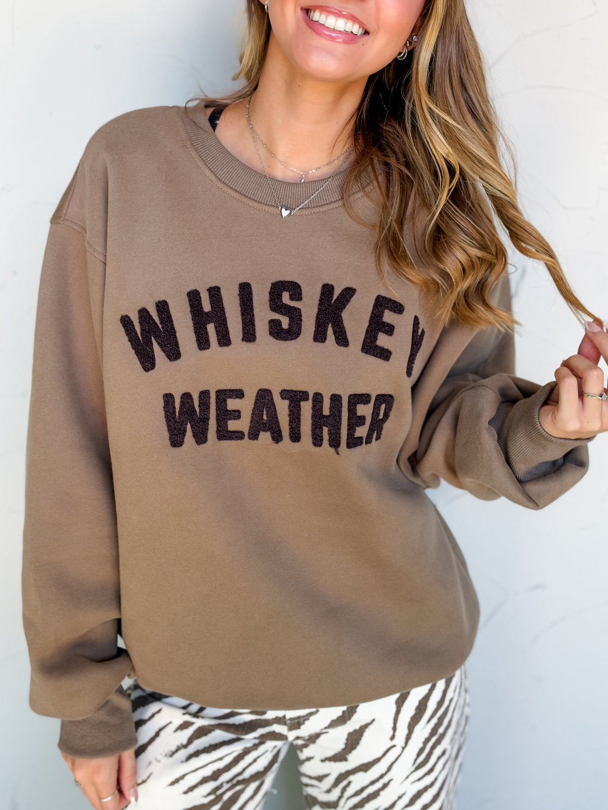 Catie wearing the Panache Apparel Co Whiskey Weather Sweatshirt in taupe brown, made of 50% cotton and 50% polyester, featuring a plain back, dropped shoulders, long sleeves, and ribbed trim, paired with cream and brown zebra-print pants.