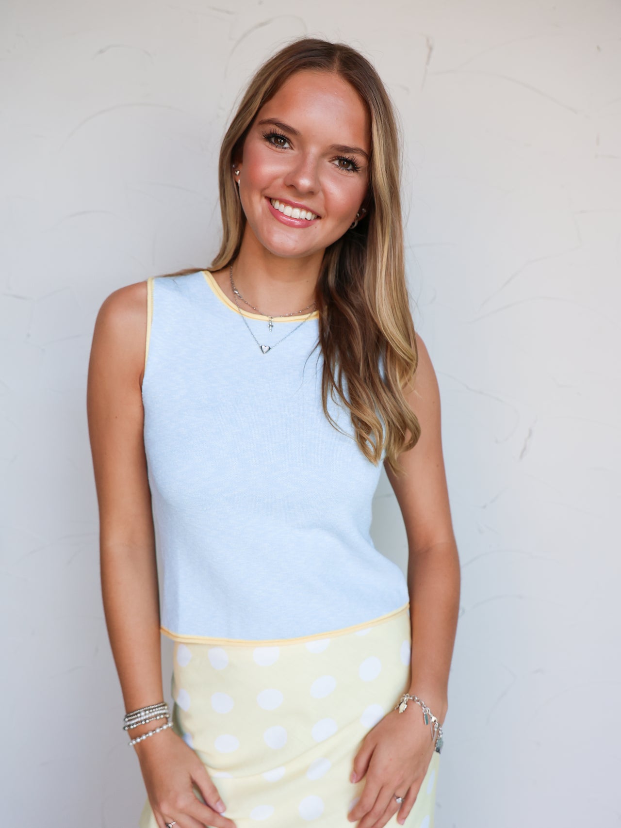 A mid-shot photograph of a smiling blonde woman posing against a textured white wall. She is wearing a fitted sleeveless light blue tank top made from a 70% viscose and 30% polyester blend fine knit fabric, featuring soft yellow contrast trim around the neckline, arm openings, and bottom hem, styled over a pastel yellow skirt with white polka dots.