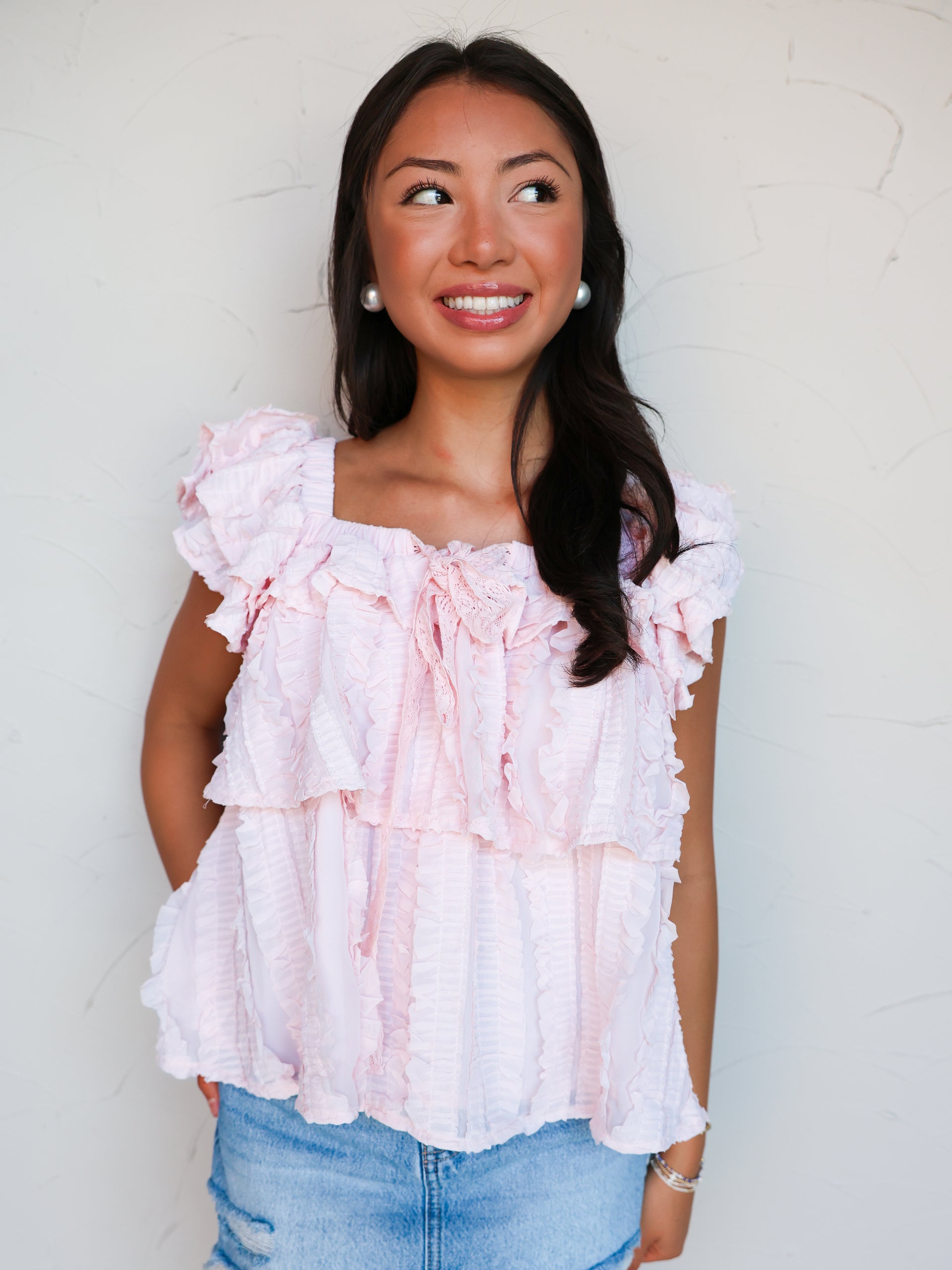 A mid-shot photograph of a smiling brunette woman looking off-camera and posing against a textured white wall. She is wearing a light pink sleeveless top made from a 100% cotton fabric featuring textured stripes, a square neckline, all-over tiered ruffle layers, and a delicate bow tie detail at the chest, styled over distressed light-wash blue denim.