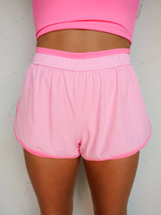 A close-up photograph focusing on the midsection of a woman posing against a textured white wall. She is wearing high-waisted athletic running shorts made from an 86% nylon and 14% spandex blend fabric featuring a light pink outer layer and a bright bubblegum pink inner lining visible at the waistband and curved dolphin hems.