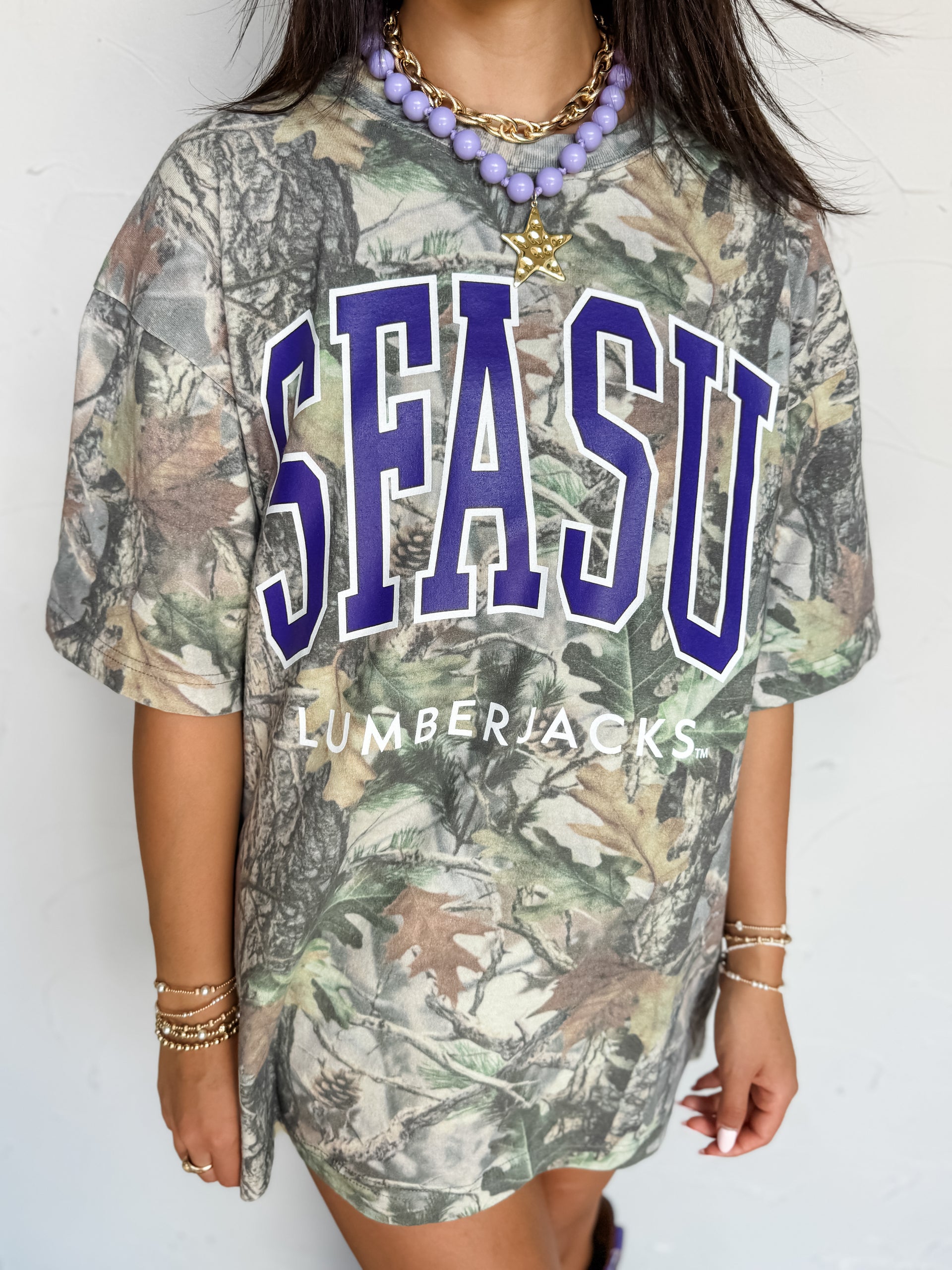 A woman wearing an oversized short-sleeve t-shirt with a woodland camouflage pattern and 'SFASU LUMBERJACKS' printed on the front in purple and white.