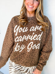Catie modeling the G Ink Carried By God Leopard Sweatshirt in a Medium, featuring brown leopard print, cream script reading “You are carried by God,” a crew neckline, and long sleeves, paired with cream and brown zebra-print pants.