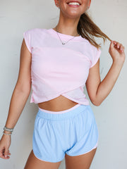 A close-up photograph from the chin down of a blonde woman posing against a textured white wall while touching her hair. She is wearing a fitted light pink activewear top made from a 65% cotton and 35% polyester blend fabric featuring short cap sleeves, a crew neckline, and a crossover tulip hemline that exposes a small triangular section of her midriff, styled over light blue athletic shorts with pink trim.