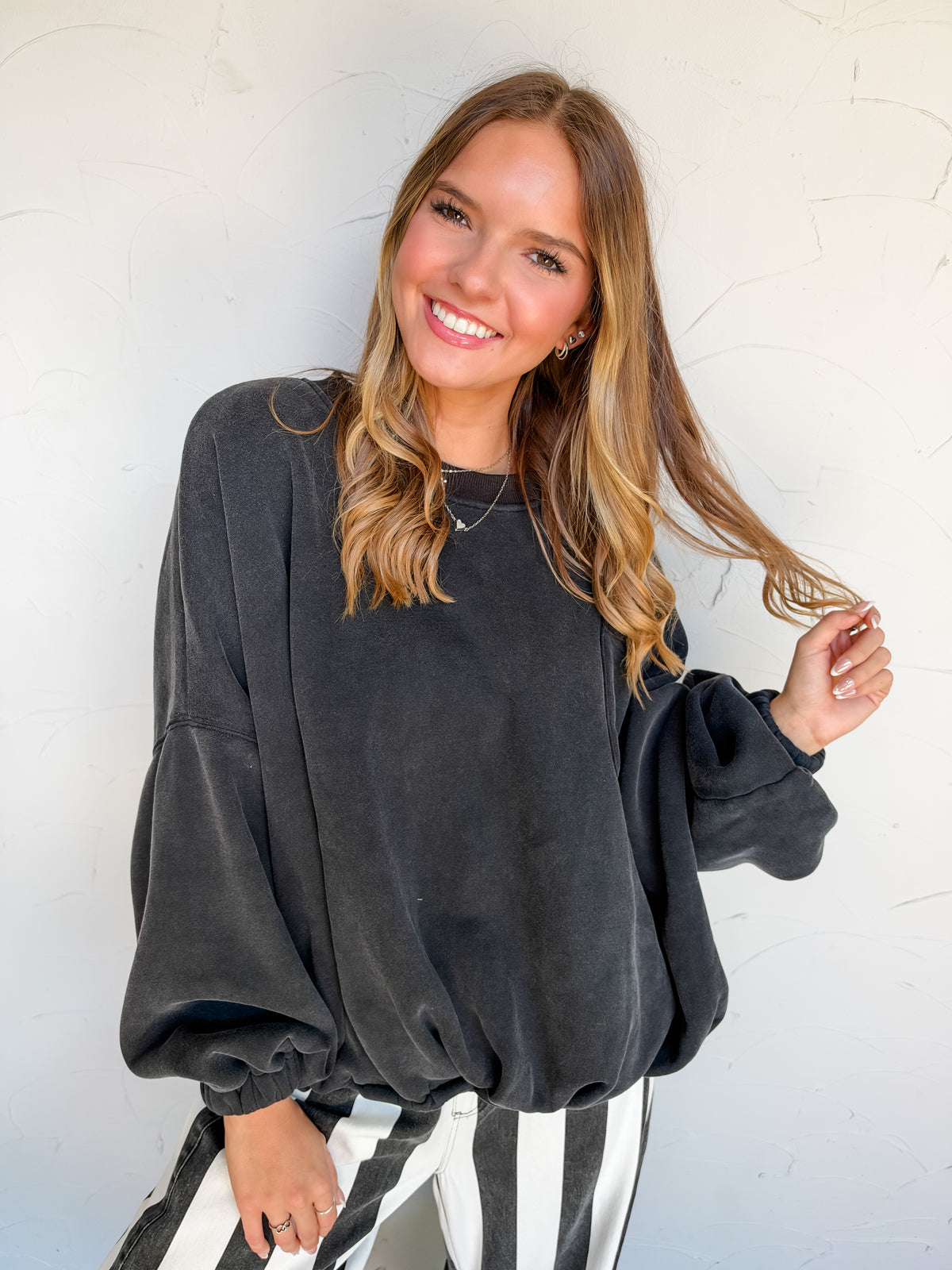 Catie wearing the LovLov Game Changer Cinch Waist Pullover in Charcoal Black, made of 50% cotton and 50% polyester, featuring dropped shoulders, long sleeves with gathered cuffs, a center-back seam, and a gathered hem, paired with black-and-white striped pants.