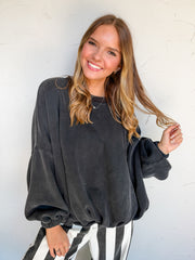 Catie wearing the LovLov Game Changer Cinch Waist Pullover in Charcoal Black, made of 50% cotton and 50% polyester, featuring dropped shoulders, long sleeves with gathered cuffs, a center-back seam, and a gathered hem, paired with black-and-white striped pants.