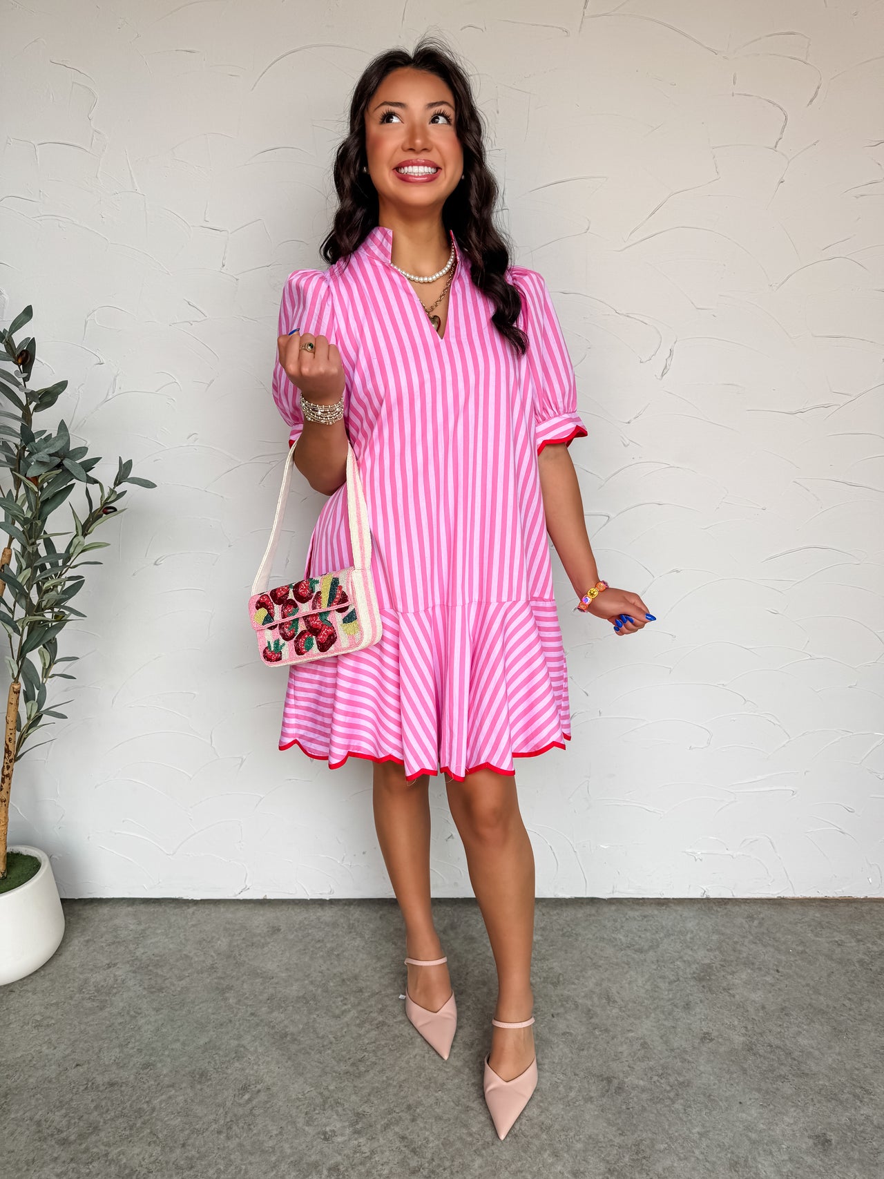 A full-length photograph of a smiling brunette woman looking upward and posing against a textured white wall while carrying a beaded strawberry-patterned shoulder bag. She is wearing a short-sleeve dress made from 100% cotton fabric featuring pink and white vertical stripes, designed with a split V-neck collar, short puff sleeves, and a dropped ruffle skirt with a contrasting crimson red trim, styled with light pink pointed-toe flats.