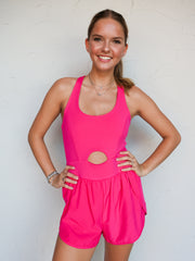 A mid-shot photograph of a smiling blonde woman posing with her hands on her hips against a textured white wall. She is wearing a hot pink one-piece activewear romper made from a 76% nylon and 24% spandex blend fabric, featuring tank straps, a scoop neckline, a small circular front cutout at the midriff, and attached running shorts with dolphin hemlines.