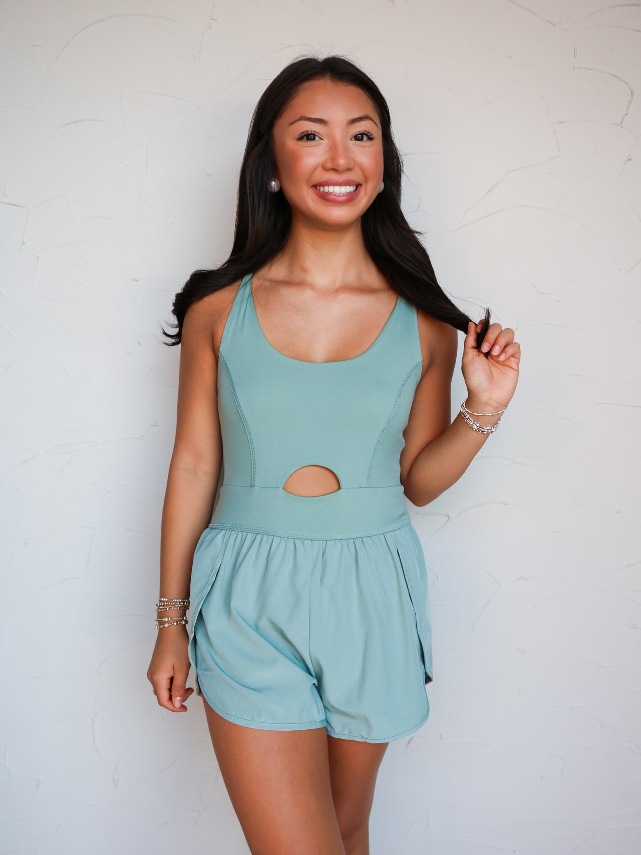 A mid-shot photograph of a smiling brunette woman posing while holding a strand of her hair against a textured white wall. She is wearing a sage green one-piece activewear romper made from a 76% nylon and 24% spandex blend fabric, featuring tank straps, a scoop neckline, a small circular front cutout at the midriff, and attached running shorts with dolphin hemlines